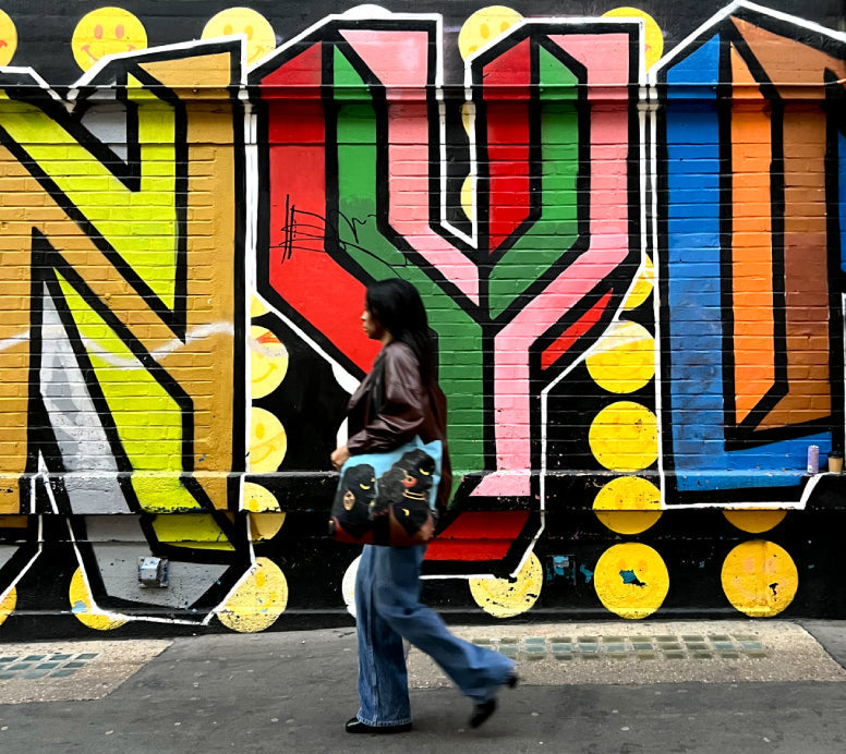 Person with a tote bag walking past a colorful mural with 'NYU' letters on a wall.